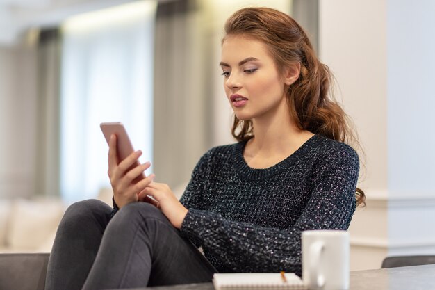 Mujer joven atractiva que mira su teléfono inteligente en casa. Mensaje de tipos de mujer en su teléfono inteligente.