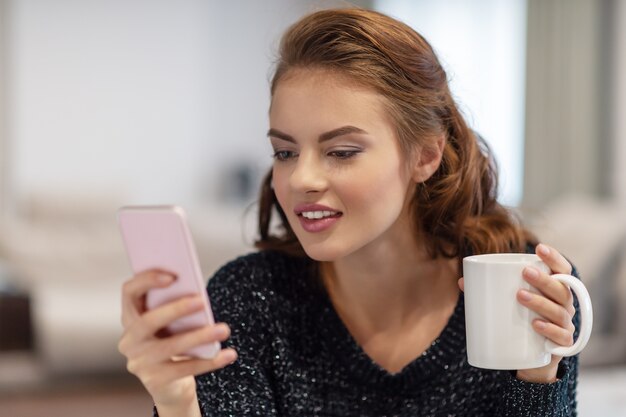 Mujer joven atractiva que mira su teléfono inteligente con una taza de café. Mensaje de tipos de mujer en su teléfono inteligente.