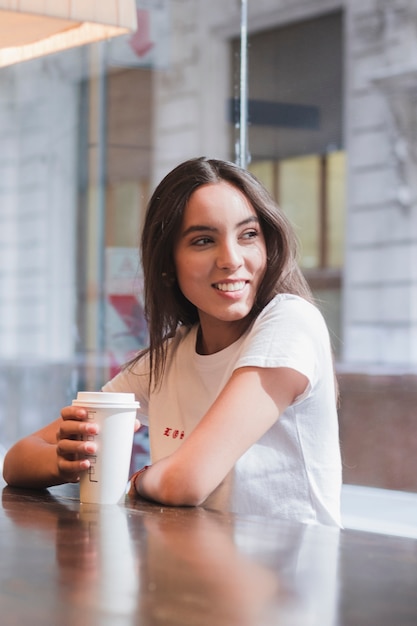 Mujer joven atractiva que se sienta en el café que sostiene la taza de café para llevar