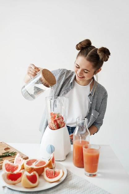 Mujer joven atractiva que sonríe agregando agua en licuadora con los pedazos de pomelo y el romero. Dieta saludable alimentación nutrición.