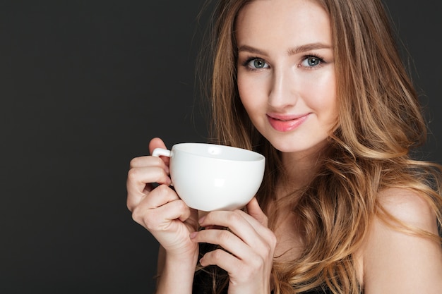 Mujer joven atractiva sonriente que sostiene la taza blanca y que bebe el café