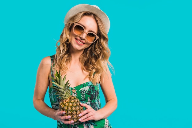 Foto gratuita mujer joven atractiva sonriente en vestido con el sombrero y las gafas de sol que sostienen la piña fresca