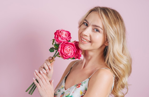 Mujer joven atractiva tímida que sostiene rosas en la mano contra el contexto rosado