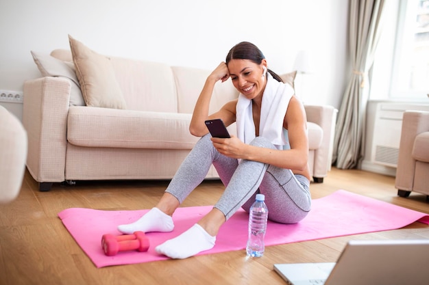 Mujer joven atractiva tomando un descanso mientras hace ejercicio en casa Mujer usando un teléfono inteligente en una colchoneta de ejercicios frente a su computadora portátil