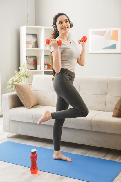 Mujer joven con auriculares haciendo ejercicio con mancuernas en estera de yoga en el sofá delantero en la sala de estar