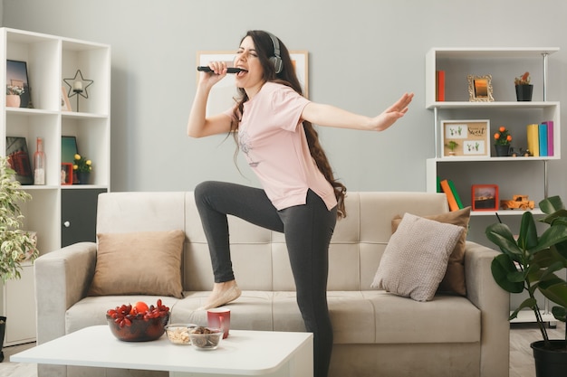 Mujer joven con auriculares con micrófono canta de pie en el sofá detrás de la mesa de café en la sala de estar