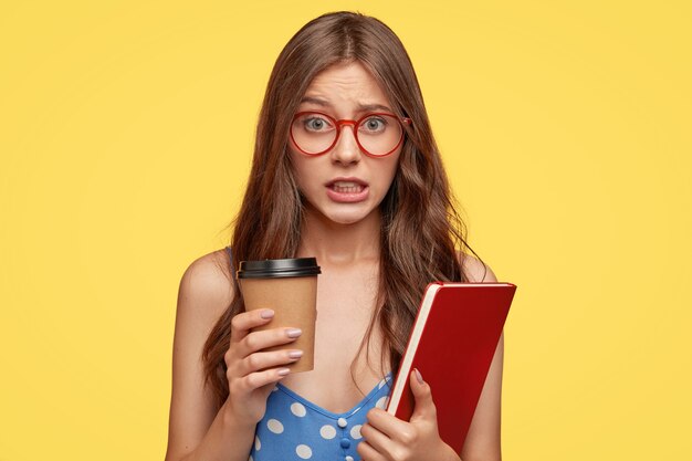 Mujer joven avergonzada con gafas posando contra la pared amarilla