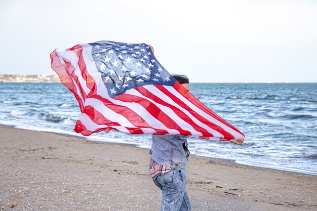 Una mujer joven con una bandera estadounidense corre junto al mar. El concepto de patriotismo y celebraciones del día de la independencia.