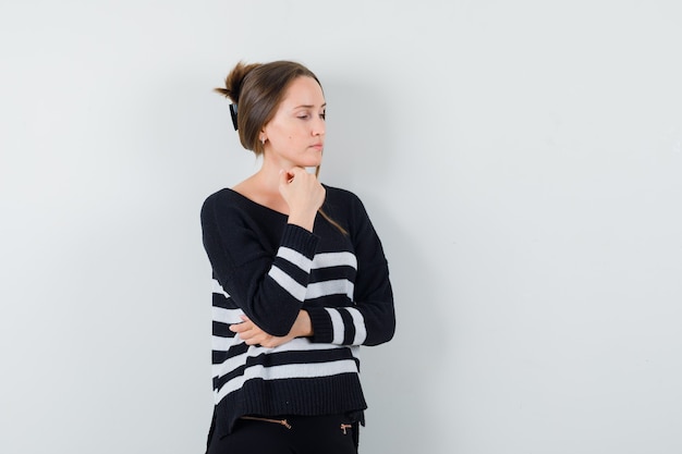 Mujer joven con la barbilla apoyada en la palma y de pie en pose de pensamiento en prendas de punto a rayas y pantalones negros y mirando pensativo