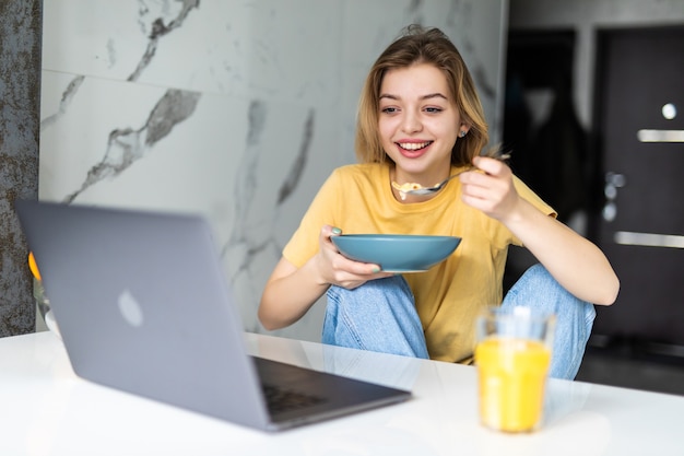 Mujer joven bastante alegre desayunando mientras está sentado en la mesa de la cocina, viendo los medios de comunicación en la computadora portátil