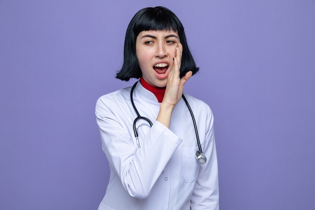 Mujer joven bastante caucásica molesta en uniforme médico con estetoscopio manteniendo la mano cerca de la boca y mirando a un lado