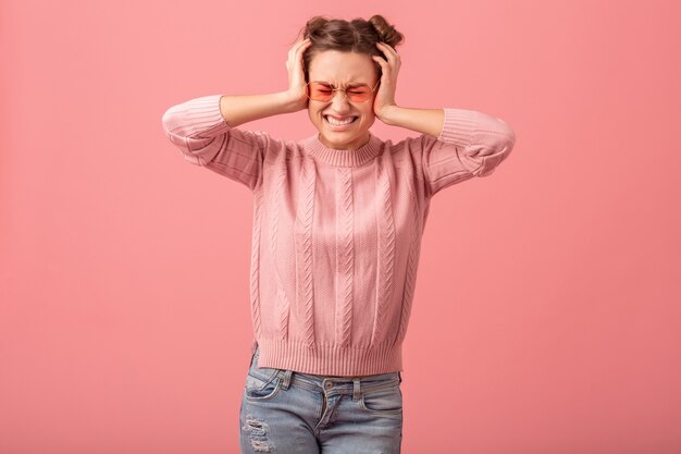 Mujer joven bastante divertida que tiene un problema, que siente dolor de cabeza, estrés y que se sostiene la cabeza, en un suéter rosa y gafas de sol aisladas sobre fondo rosa de estudio