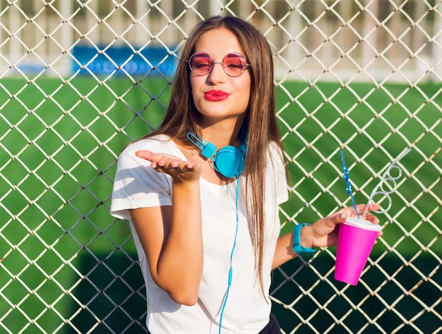 Mujer joven bastante encantadora hipster posando con taza de jugo saludable, escuchando música favorita