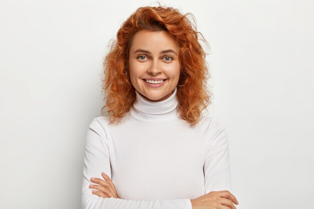 Mujer joven bastante encantadora con el pelo corto de jengibre, tiene dientes blancos perfectos y piel limpia y saludable, mantiene las manos cruzadas, recibe buenas noticias, mira directamente, aislado en la pared blanca