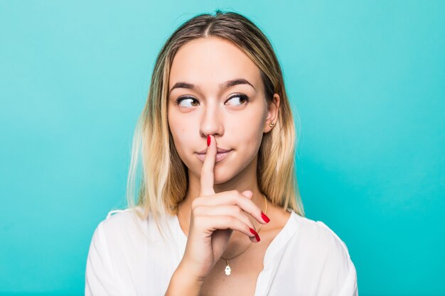 Mujer joven bastante encantadora que tiene secreto mientras mantiene el dedo en los labios y muestra el signo de silencio mientras está de pie en la pared azul