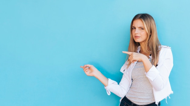 Mujer joven bastante rubia dudosa que señala los dedos al lado contra fondo azul