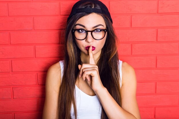 Mujer joven bastante seductora hipster divirtiéndose y mostrando shhh ciencia, vestida con gorra swag negra y gafas transparentes, cabellos largos y maquillaje brillante.