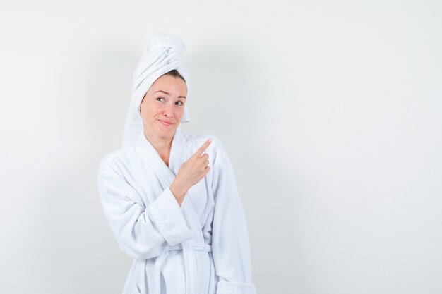 Mujer joven en bata de baño blanca, toalla apuntando a la esquina superior derecha y mirando alegre, vista frontal.