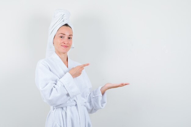 Mujer joven en bata de baño blanca, toalla apuntando a su palma extendida a un lado y mirando alegre, vista frontal.