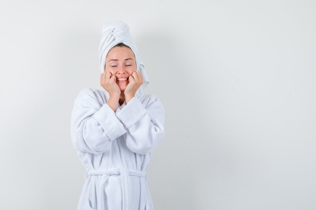 Mujer joven en bata de baño blanca, toalla manteniendo las manos en las mejillas y mirando alegre, vista frontal.