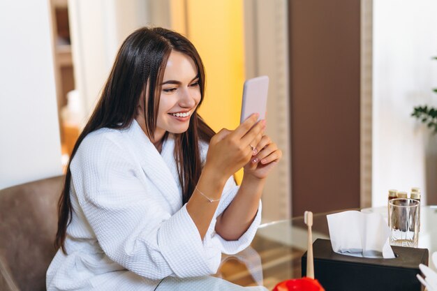 Mujer joven en bata de baño en la habitación del hotel mediante teléfono móvil