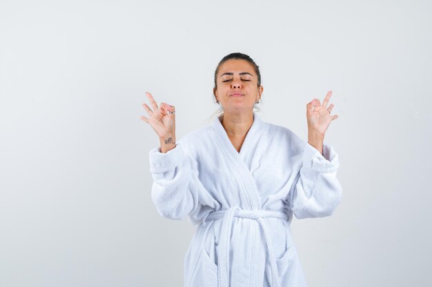 Mujer joven en bata de baño mostrando gesto de meditación y mirando pacífica