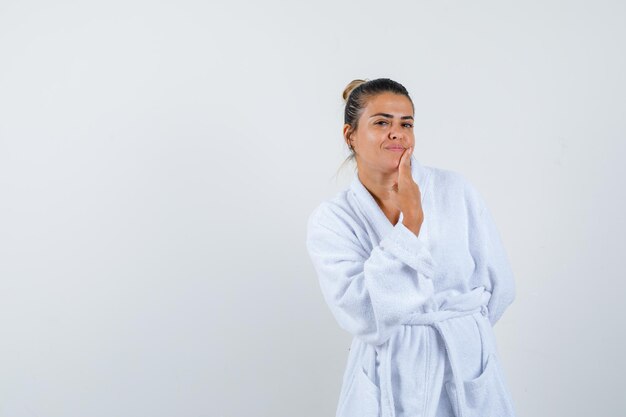 Mujer joven en bata de baño tocando la barbilla con el dedo y mirando lindo