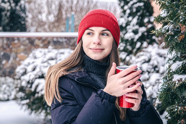 Una mujer joven bebe una bebida caliente de una taza térmica roja en invierno