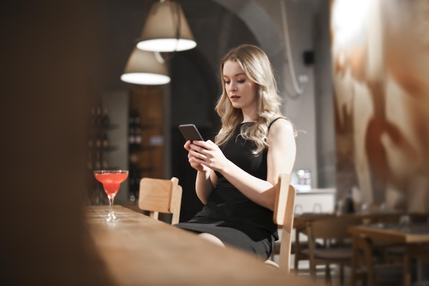 mujer joven bebe un cóctel en un bar y lee desde su teléfono inteligente