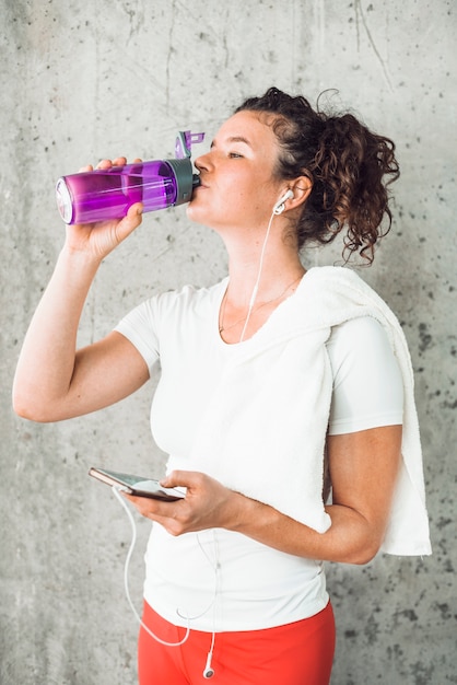 Mujer joven bebiendo agua y escuchando música en un teléfono inteligente