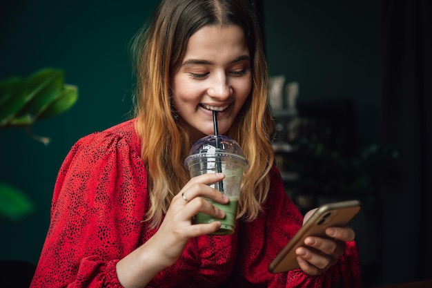 Mujer joven bebiendo bebida verde hielo matcha latte en café y usando teléfono inteligente