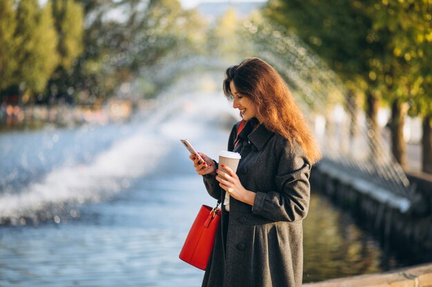 Mujer joven bebiendo café y usando el teléfono en el parque