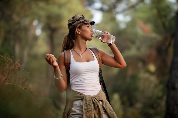 Mujer joven bebiendo haver y refrescándose mientras camina por la naturaleza