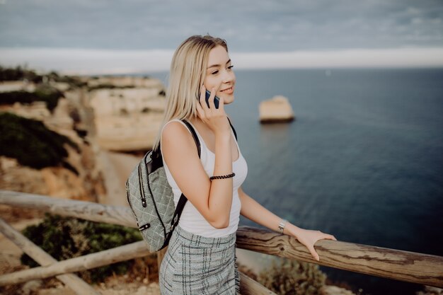 Mujer joven belleza hablando por teléfono móvil junto al océano con panorama de rocas.