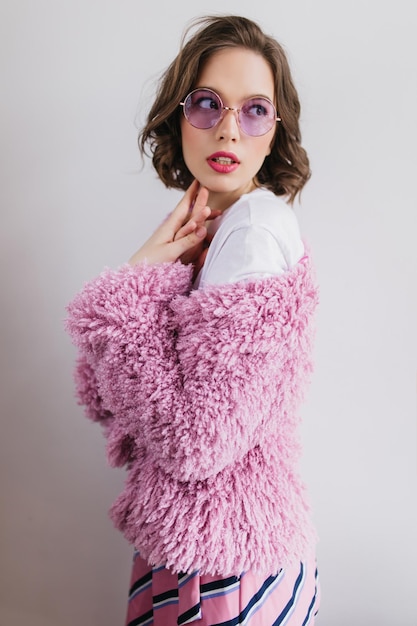 Mujer joven bien formada con cabello castaño ondulado posando en una chaqueta de piel Foto interior de una chica guapa interesada en gafas de sol y abrigo esponjoso