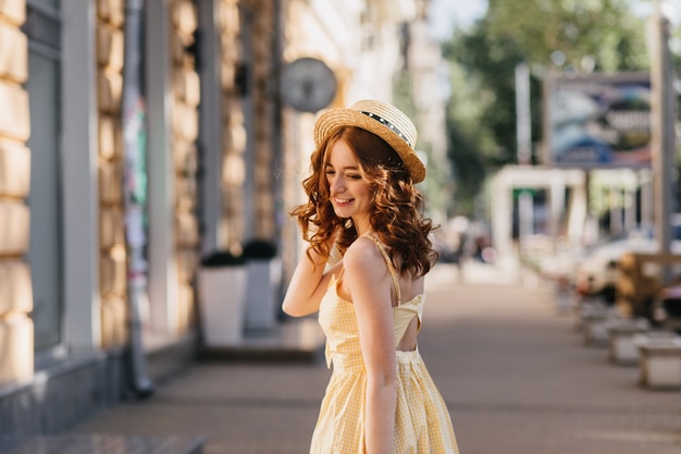 Mujer joven bien formada en vestido amarillo posando con placer en la ciudad. Foto al aire libre de la impresionante chica con sombrero disfrutando de la sesión de fotos durante la caminata.