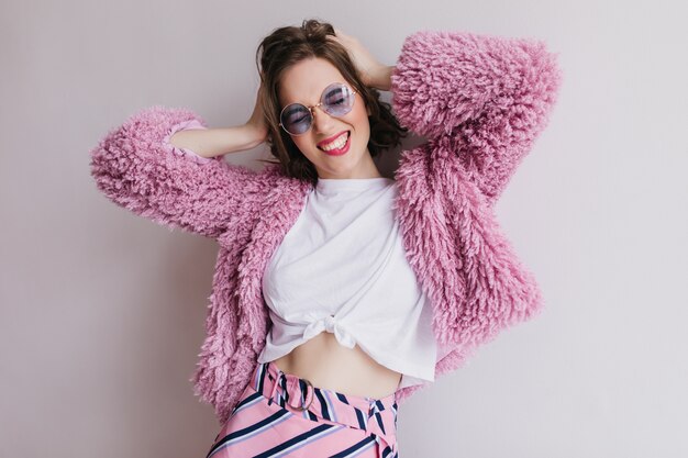 Mujer joven bien vestida emocionada que presenta en la pared ligera. atractiva chica morena en abrigo de piel rosa jugando con su cabello y riendo.