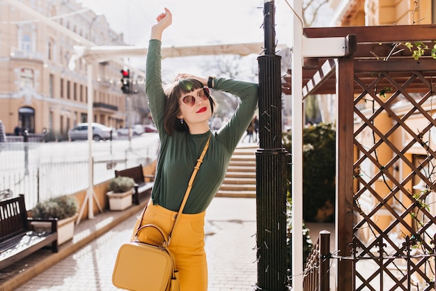 Mujer joven bien vestida posando con las manos en la ciudad. Tiro al aire libre de la dichosa chica morena con gafas de sol disfrutando del buen tiempo.