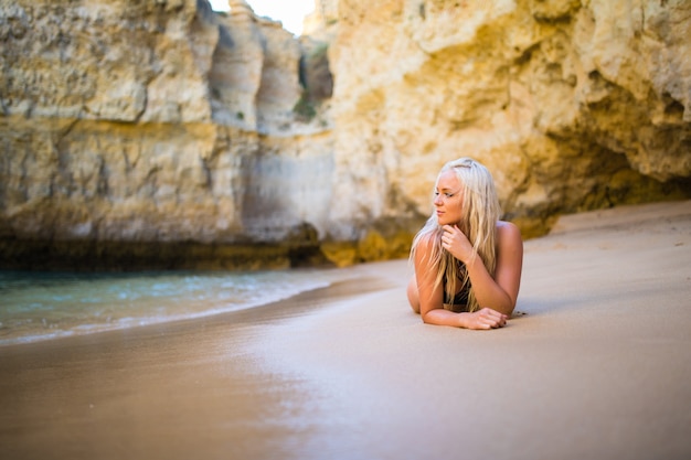 Una mujer joven en bikini negro con un esbelto cuerpo bronceado y hermosos pechos se encuentra en un banco de arena junto al agua al atardecer en septiembre, otoño siberiano