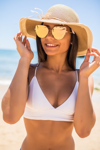 Foto gratuita mujer joven en bikini con sombrero de paja blanco disfrutando de las vacaciones de verano en la playa. retrato de hermosa mujer latina relajante en la playa con gafas de sol.