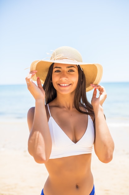 Mujer joven en bikini con sombrero de paja blanco disfrutando de las vacaciones de verano en la playa. Retrato de hermosa mujer latina relajante en la playa con gafas de sol.