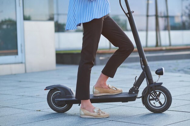 Mujer joven en blaser despojado está montando su nuevo scooter eléctrico en la calle.