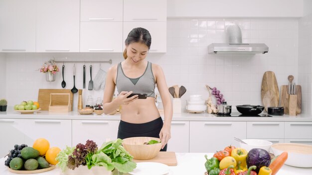 Mujer joven blogger asiática con publicación de fotos de teléfono inteligente en las redes sociales en la cocina