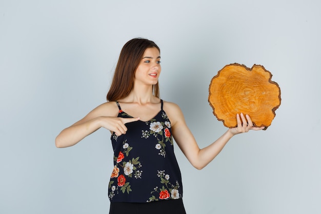 Mujer joven en blusa apuntando al trozo de madera y mirando alegre, vista frontal.