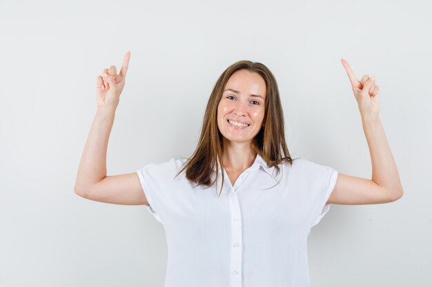 Mujer joven en blusa blanca apuntando hacia arriba y mirando feliz
