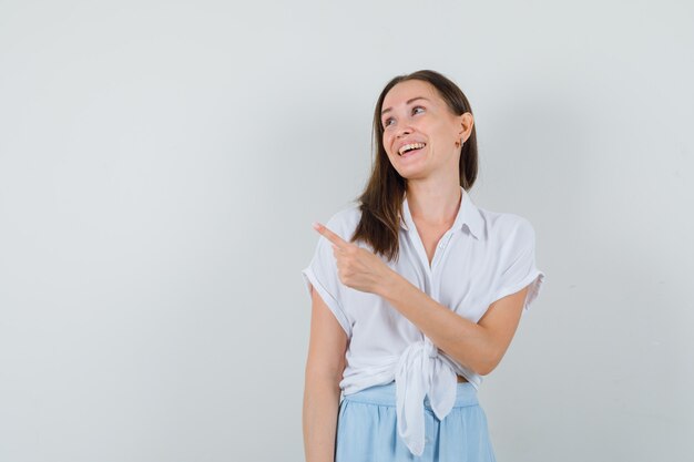 Mujer joven en blusa blanca y falda azul claro apuntando hacia la izquierda con el dedo índice y mirando alegre