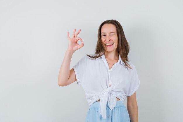 Mujer joven en blusa blanca y falda azul claro que muestra el signo bien y sacando la lengua y mirando alegre