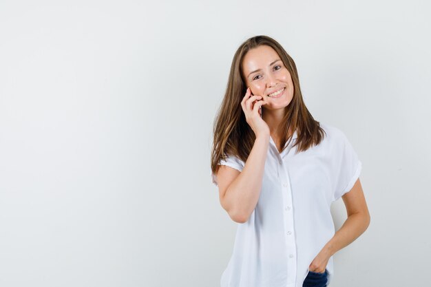 Mujer joven en blusa blanca hablando por teléfono y luciendo optimista