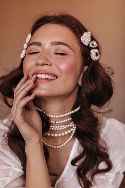 Mujer joven en blusa blanca y joyas de perlas se ríe. Instantánea de mujer con pecas sobre fondo beige.