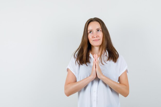 Mujer joven en blusa blanca mostrando gesto de oración y mirando enfocado